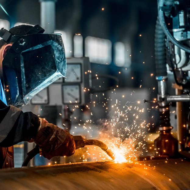 A person in blue clothing welds on metal. The room is a bit dark.