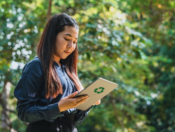 Person holding a tablet with a recycling symbol, standing outdoors surrounded by lush green trees, promoting environmental awareness.