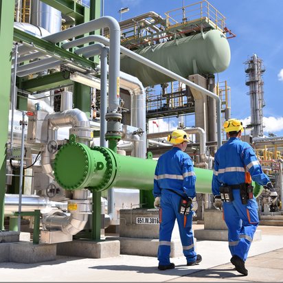Two people dressed in blue are walking around and inspecting. The inspection is on an industrial estate, there are many pipes in the background. In the right corner is a clear blue sky.