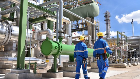 Two people dressed in blue are walking around and inspecting. The inspection is on an industrial estate, there are many pipes in the background. In the right corner is a clear blue sky.