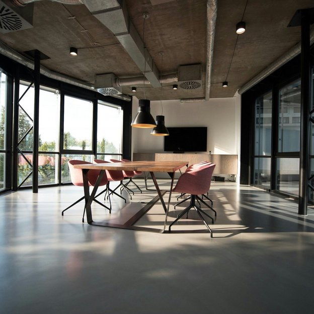 An empty, modern conference room with large windows, a wooden table, pink chairs, and hanging ceiling lights.