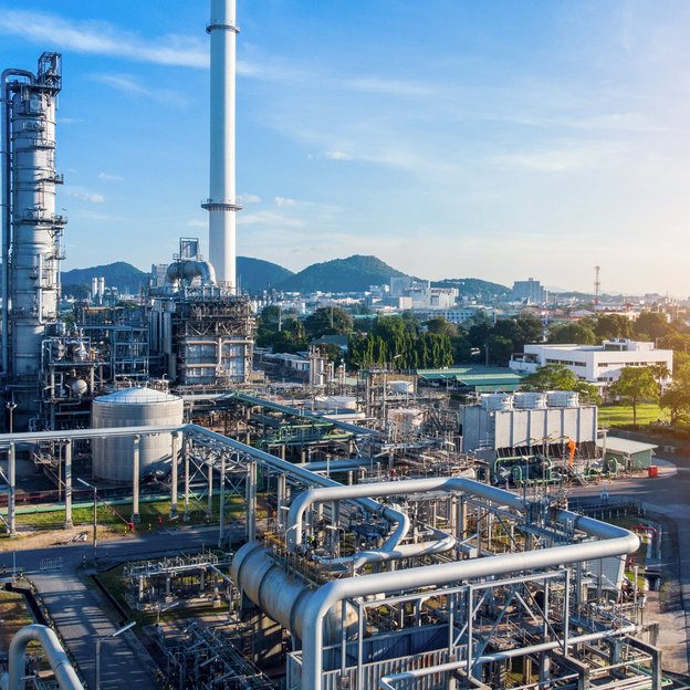 A picture of the refinery. To the right is a row of trees. In the top right corner the sun is shining.