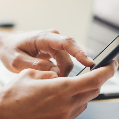 A person is sitting and pressing something on a phone, to the left of the person is a computer.