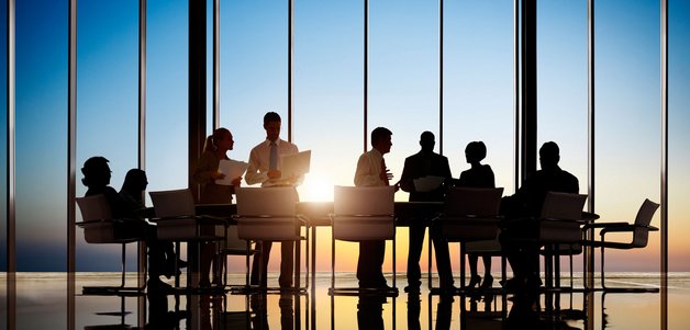 A group of people are gathered around a conference table in a modern office setting, silhouetted against large windows showing a sunset. The scene conveys a professional atmosphere with discussions taking place.