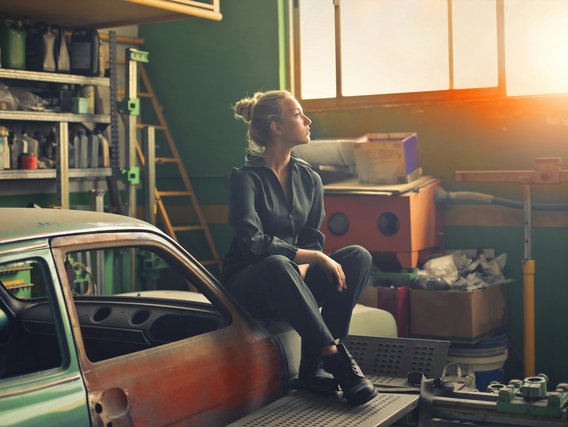 A woman in a dark jumpsuit is sitting on a car part in a workshop, gazing out of a window with sunlight streaming in.