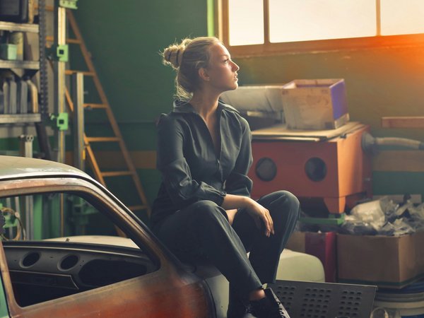 A woman in a dark jumpsuit is sitting on a car part in a workshop, gazing out of a window with sunlight streaming in.