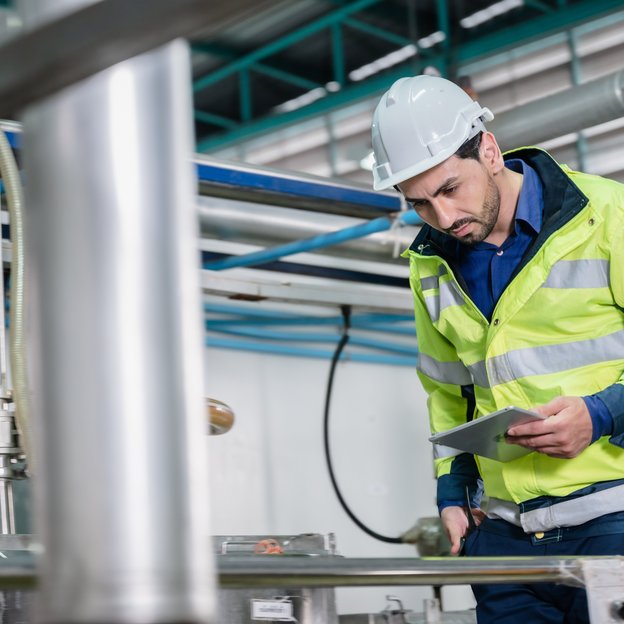 A person wearing a white helmet stands looking partly at his tablet and down at some pipes.