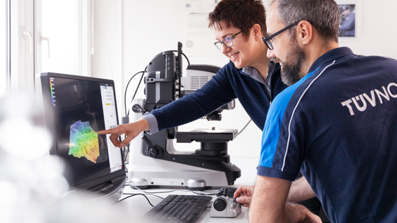 Two engineers analyze 3D data on a computer screen in a laboratory setting. The male has TÜV NORD logo on the back of his pike shirt.