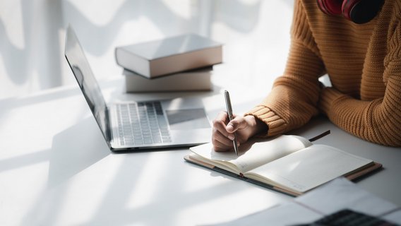 A person is writing in a book. Next to the book is the computer with the screen up. Further up in the picture, next to the computer, two books are stacked.