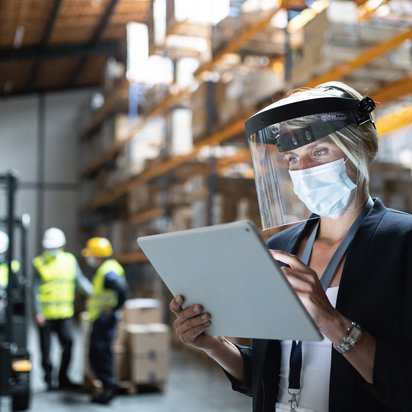 Warehouse worker using a tablet with personal protective equipment in a storage facility Person wearing protective face shield and mask using a tablet in a warehouse with shelves and forklift in the background.