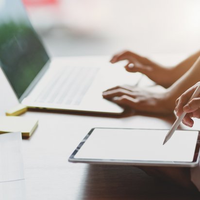 Two people are standing at a table. The person closest to the camera is holding a tablet and a pen. The other person presses the keys on a computer.