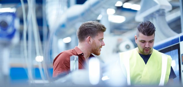 Two individuals are engaged in conversation in an industrial setting, surrounded by machinery and equipment. One person is wearing a high-visibility vest, indicating a focus on safety in the workplace.