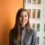Smiling Woman Sitting by a Window with a Laptop A young woman with long brown hair, wearing a gray blazer, sits by a window holding a laptop on her lap. She is smiling, and the background shows the exterior of a building through the window.