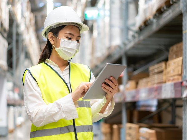 Worker in high-visibility vest inside a warehouse and hard hat using a tablet, surrounded by shelves filled with boxes and packages.