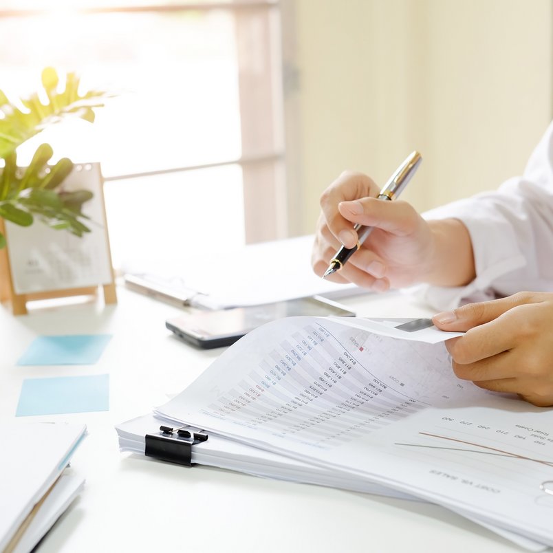 A person is sitting at his/her desk, holding a pen and leafing through some documents. On the desk there is also a plant and some notes.
