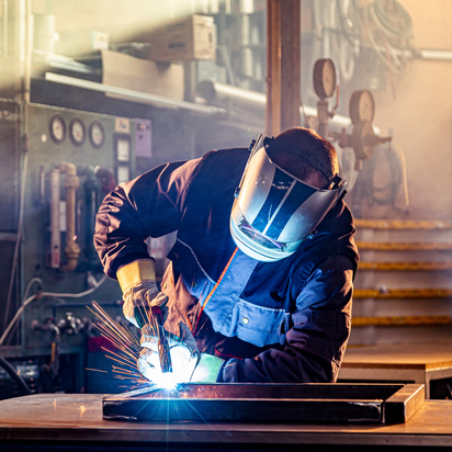 A person is bend over a steel structure and welding at parts of square steel lying on a table.