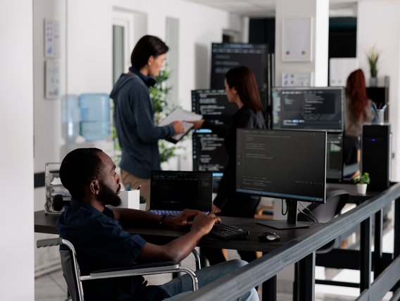 Modern office environment with personnel working on computers, discussing tasks, and collaborating on projects amidst a tech-focused setup.