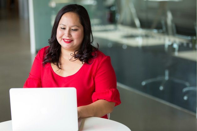 A woman with dark hair, wearing a bright red blouse, sits at a white table using a laptop. She is smiling as she works in a modern office setting.