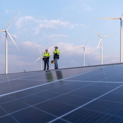 Inspektion von Photovoltaik- und Windenergieanlagen durch TÜV-Fachkräfte Zwei TÜV NORD-Ingenieure prüfen Solarmodule vor Windrädern unter blauem Himmel