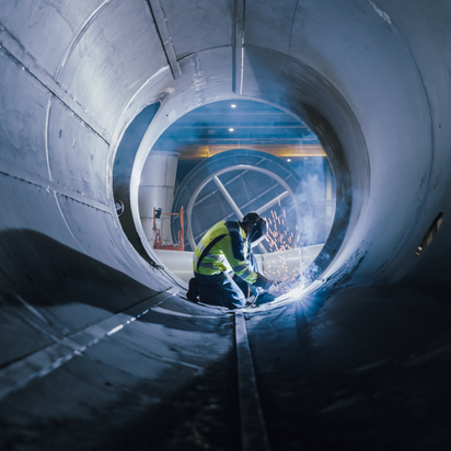 A welder works inside a large metal pipe, creating sparks in an industrial setting.