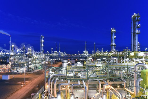 A refinery is seen, to the left there is a road. The sky is darkblue, light comes from the refinery.