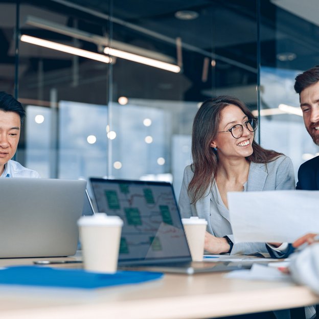 Group of professionals collaborating in a sleek, glass-walled office. Laptops, documents, and coffee cups are on the table, indicating a productive meeting.