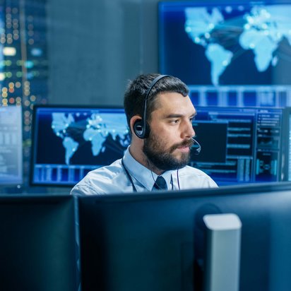 Monitoring data and global systems in a control room environment Person wearing a headset working at multiple computer monitors displaying data and world maps in a control room