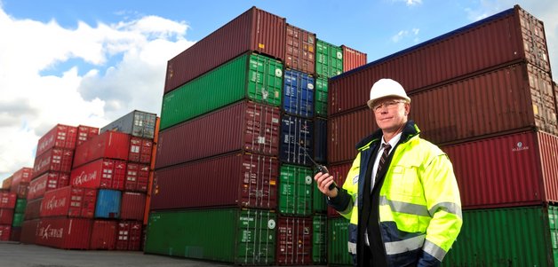 A person in a high-visibility jacket and hard hat stands in front of stacked shipping containers, holding a walkie-talkie. The scene is set outdoors under a blue sky with scattered clouds, indicating a busy logistics or shipping environment.