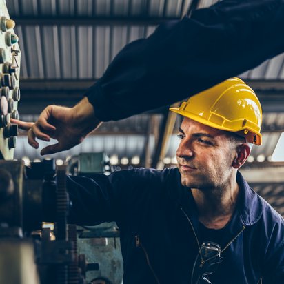 Two people are standing in a factory. The nearest one presses a button. The other person stands and looks towards the button being pressed.