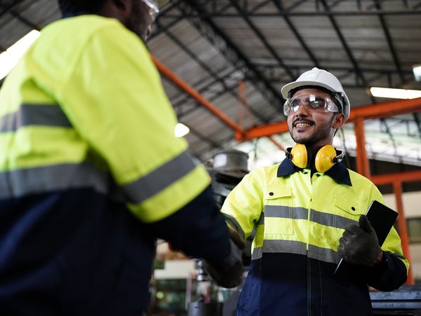 Two workers in high-visibility jackets and hard hats shaking hands in an industrial setting, emphasizing teamwork and safety.