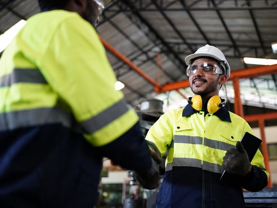 Two workers in high-visibility jackets and hard hats shaking hands in an industrial setting, emphasizing teamwork and safety.