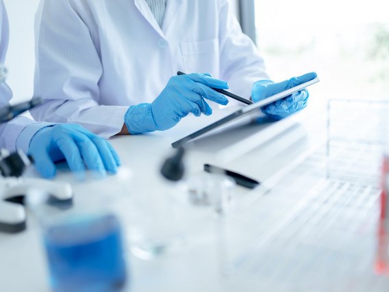 Two scientists wearing protective gloves are documenting data on a tablet while working in a laboratory, possibly during an experiment or analysis.